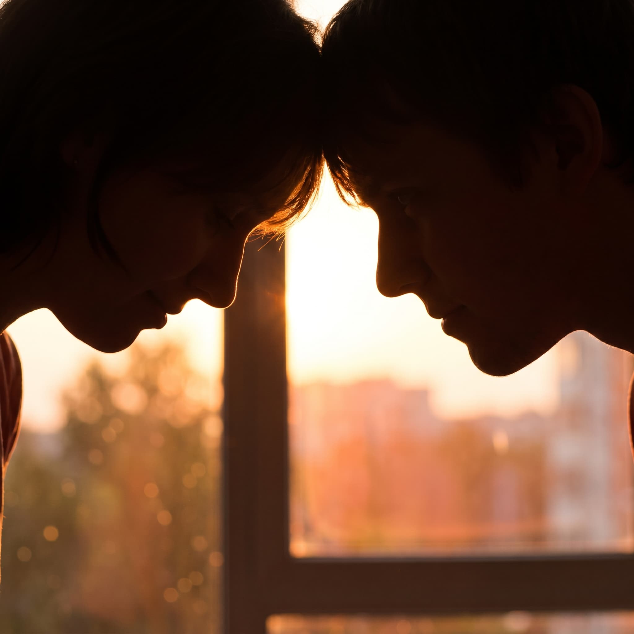 Two silhouettes with foreheads almost touching against a golden window backlight.
